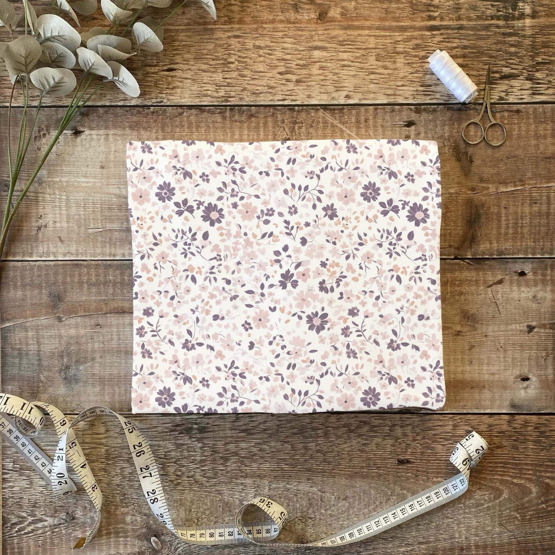 Ein Stück Stoff mit feinem Blumenmuster in Lila und Rosa liegt auf einem rustikalen Holztisch und zeigt die Größe und Wirkung des Designs.